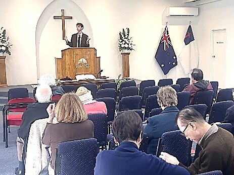 John Cirone preaching in an Australian church, the national flag behind him.